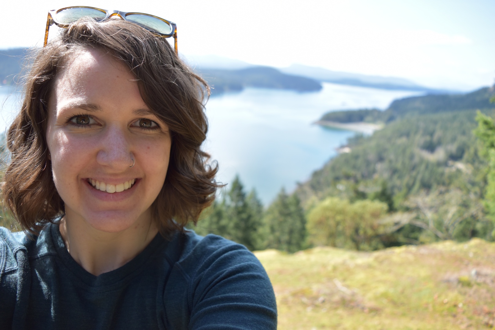 Kaitlyn is smiling at the camera with sunglasses on top of her head. Over her left shoulder are tree covered islands with a fjord cutting through them.